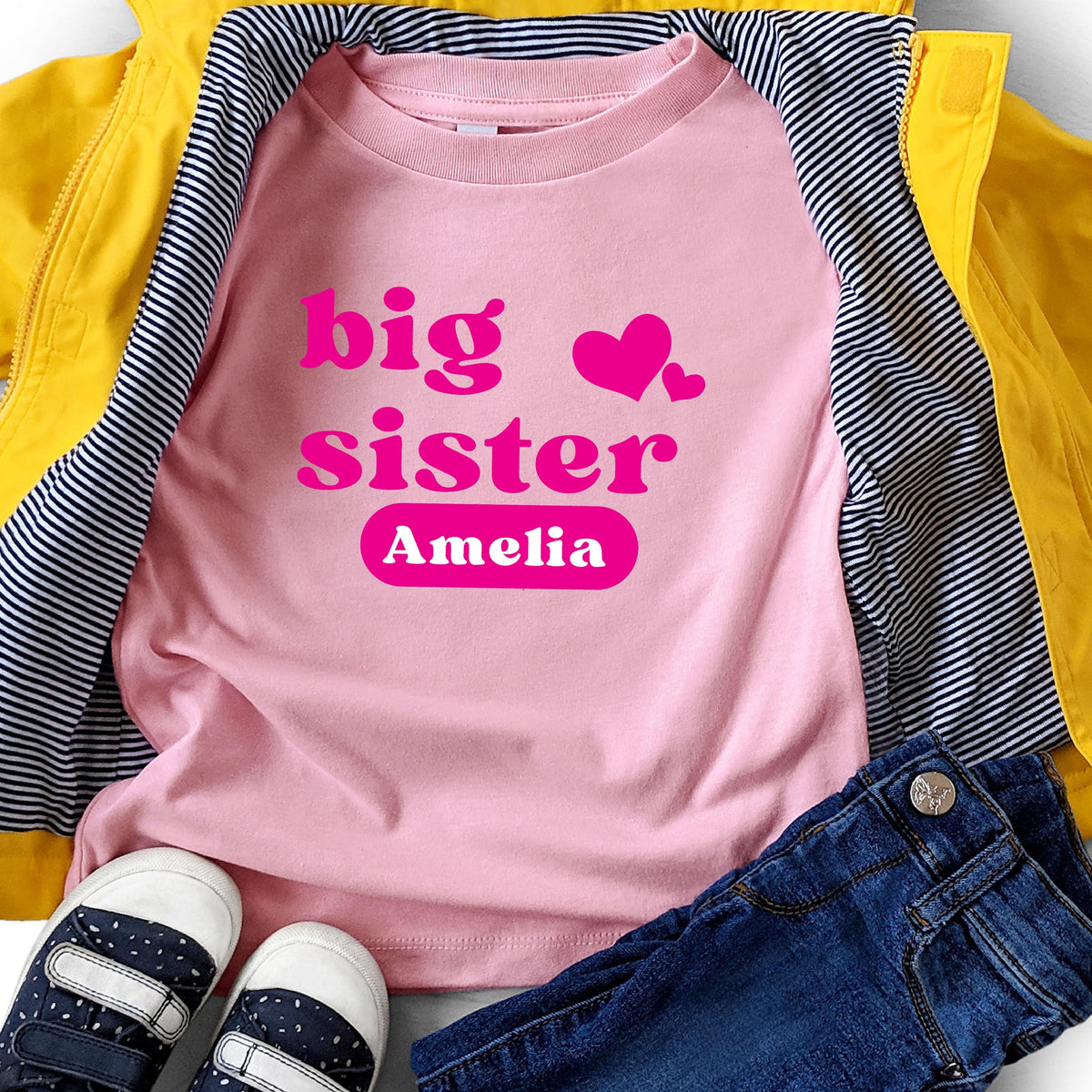 May include: A light pink t-shirt featuring the hot pink text big sister and two hearts, with the name Amelia printed in white inside a hot pink rounded rectangle. The shirt is layered under a bright yellow jacket and dark blue denim jeans.