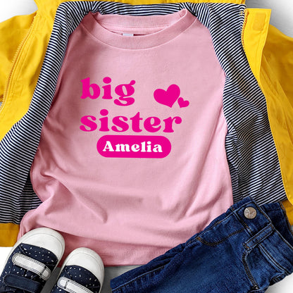 May include: A light pink t-shirt featuring the hot pink text big sister and two hearts, with the name Amelia printed in white inside a hot pink rounded rectangle. The shirt is layered under a bright yellow jacket and dark blue denim jeans.