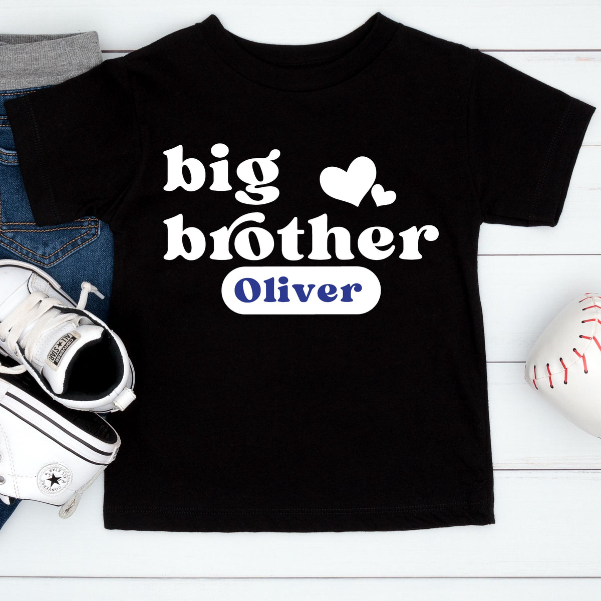 May include: A black short-sleeved t-shirt featuring the white text "big brother" with two white hearts above it. Below the text is a white rounded rectangular patch containing the name "Oliver" printed in blue capital letters.