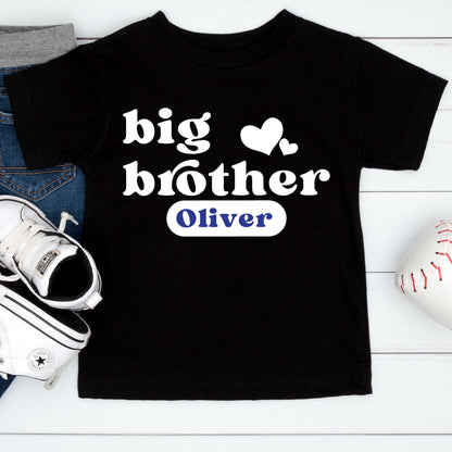 May include: A black short-sleeved t-shirt featuring the white text "big brother" with two white hearts above it. Below the text is a white rounded rectangular patch containing the name "Oliver" printed in blue capital letters.