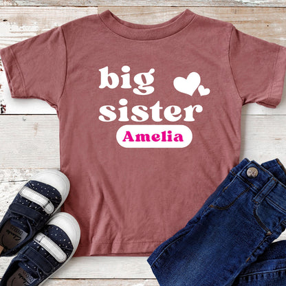 May include: A dusty rose short-sleeved t-shirt featuring the white text big sister with two white hearts and the name Amelia printed in pink inside a white rounded rectangle, accompanied by dark blue denim jeans and navy blue sneakers.
