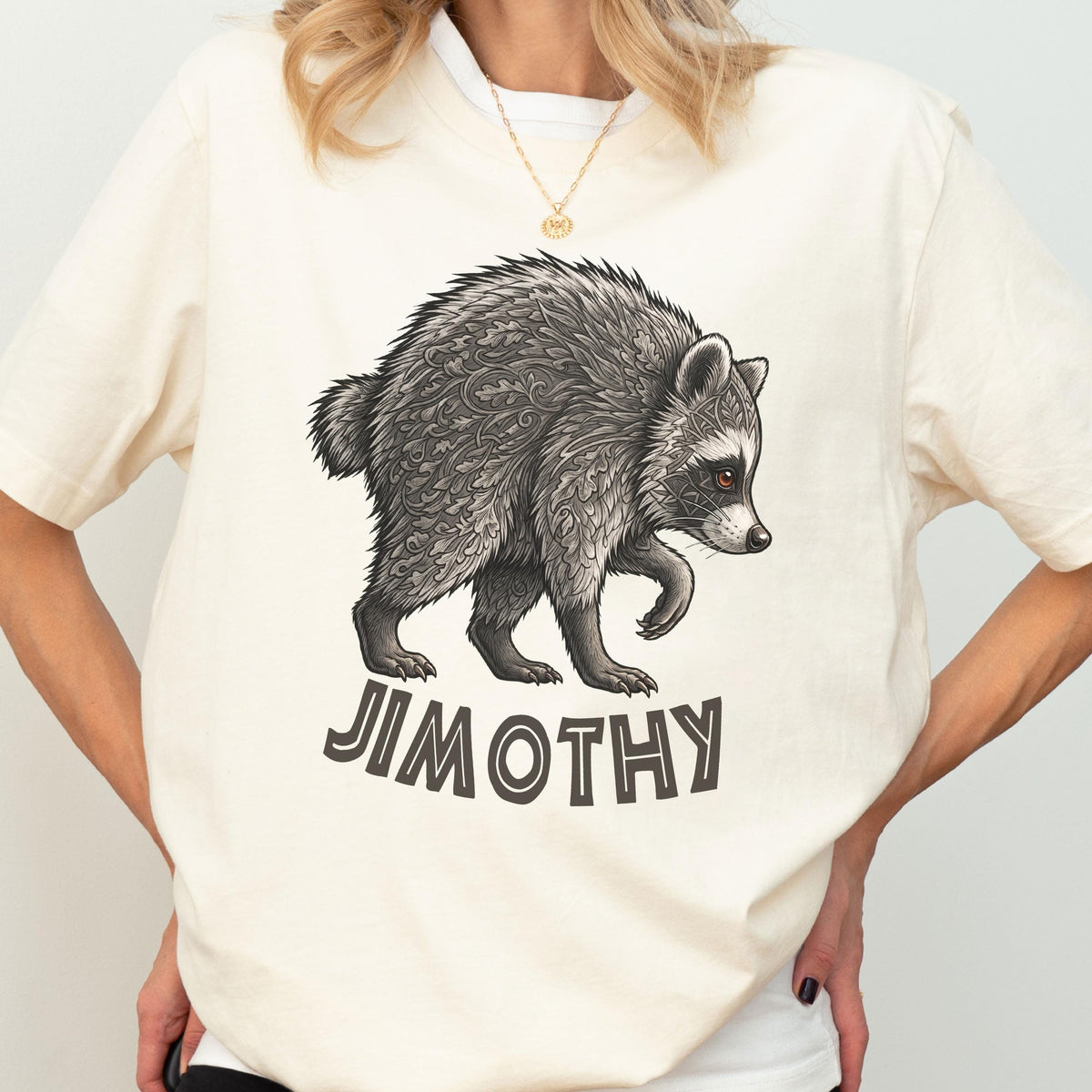 May include: A cream-colored short-sleeve t-shirt featuring a detailed illustration of a raccoon with intricate leaf-like patterns on its fur. Below the raccoon, the text JIMOTHY is printed in a bold, dark, sans-serif font on the front center.