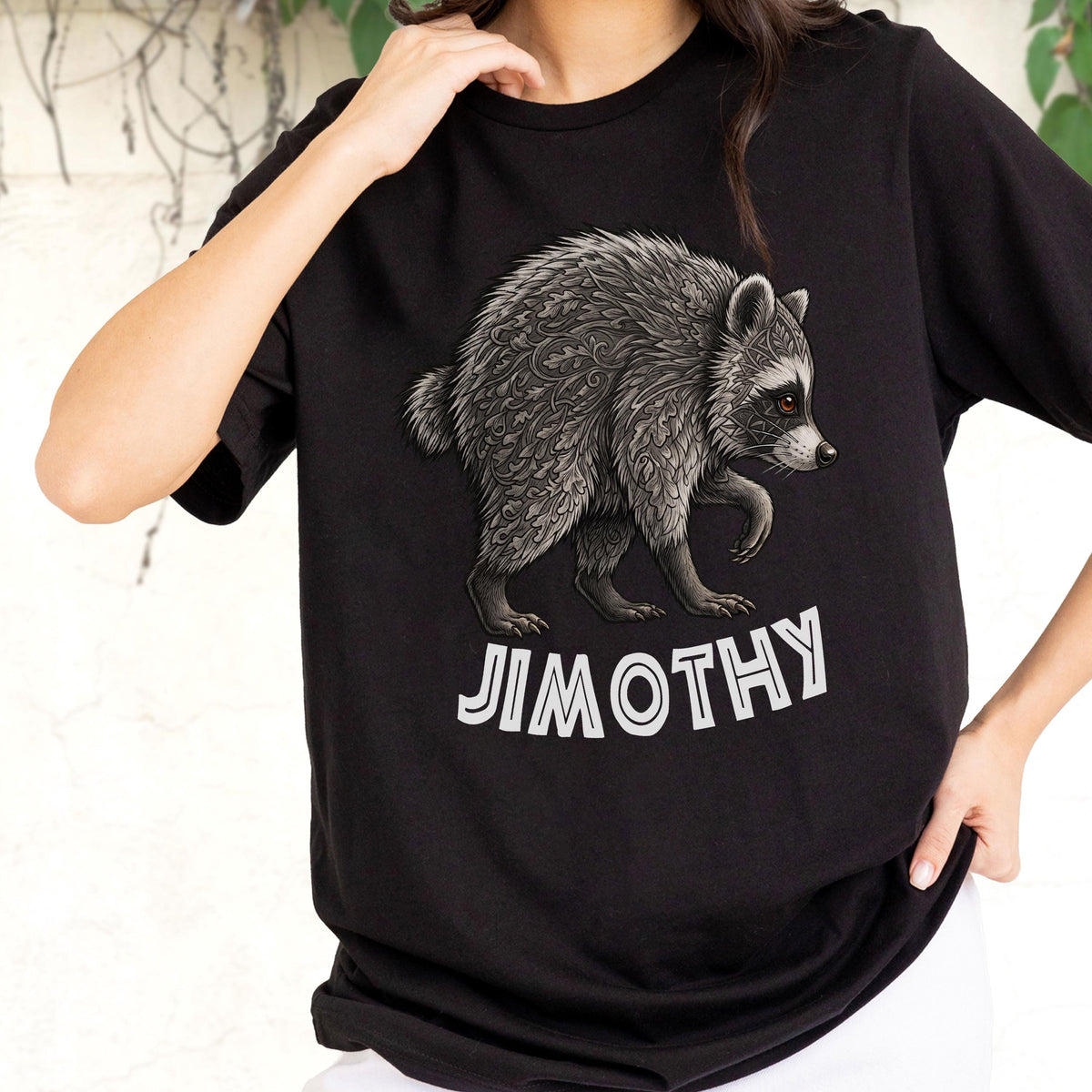 May include: A black short-sleeved t-shirt featuring a graphic of a raccoon with intricate leaf-like patterns on its fur. Below the raccoon, the name JIMOTHY is printed in bold, white, sans-serif capital letters. The shirt has a crew neckline.