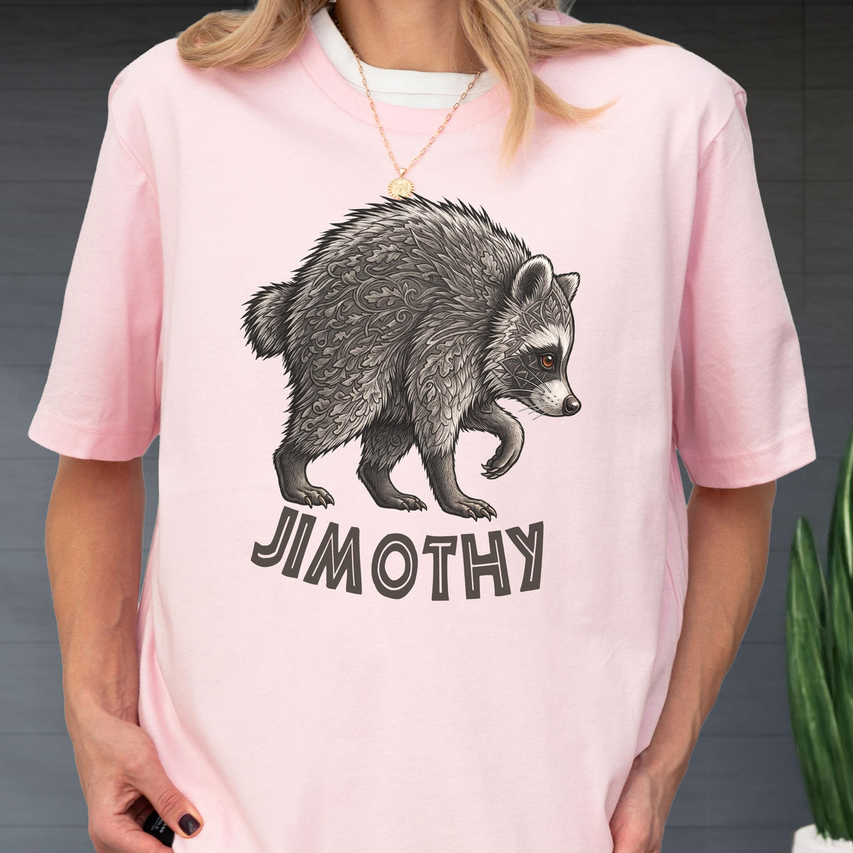 May include: A light pink short-sleeve t-shirt featuring a detailed graphic of a raccoon with intricate leaf-like patterns on its fur. Below the raccoon, the text JIMOTHY is printed in a bold, dark gray, sans-serif font on the front of the shirt.
