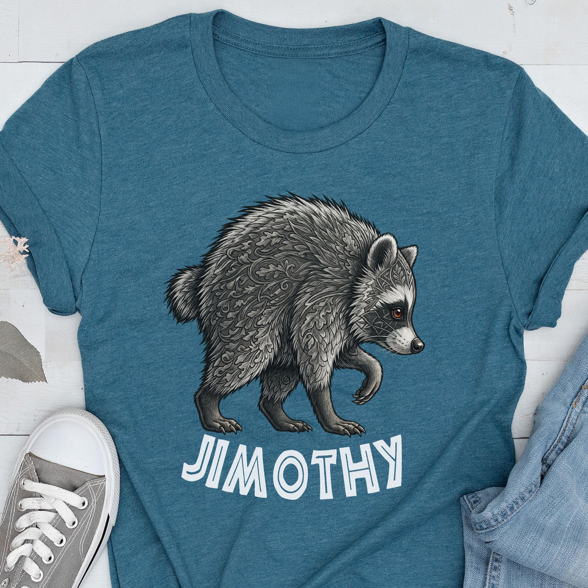 May include: A teal blue t-shirt featuring a detailed graphic of a raccoon with intricate leaf-like patterns in its fur. Below the raccoon, the name JIMOTHY is printed in bold, white, capital letters. The shirt has a crew neckline and short sleeves.