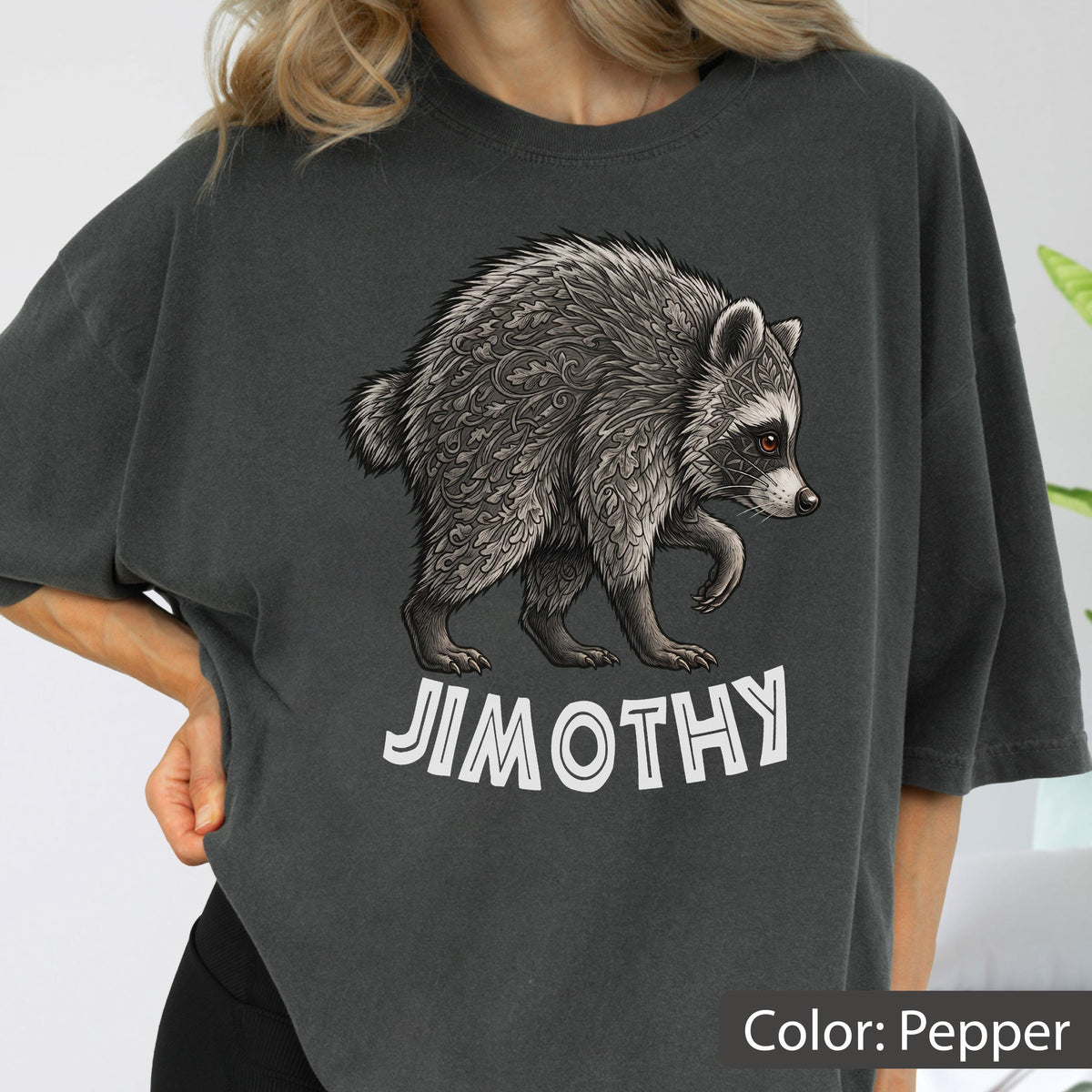 May include: A charcoal gray t-shirt featuring a graphic illustration of a raccoon with intricate, swirling leaf-like patterns throughout its fur. Below the raccoon, the name JIMOTHY is printed in bold, white, sans-serif capital letters.