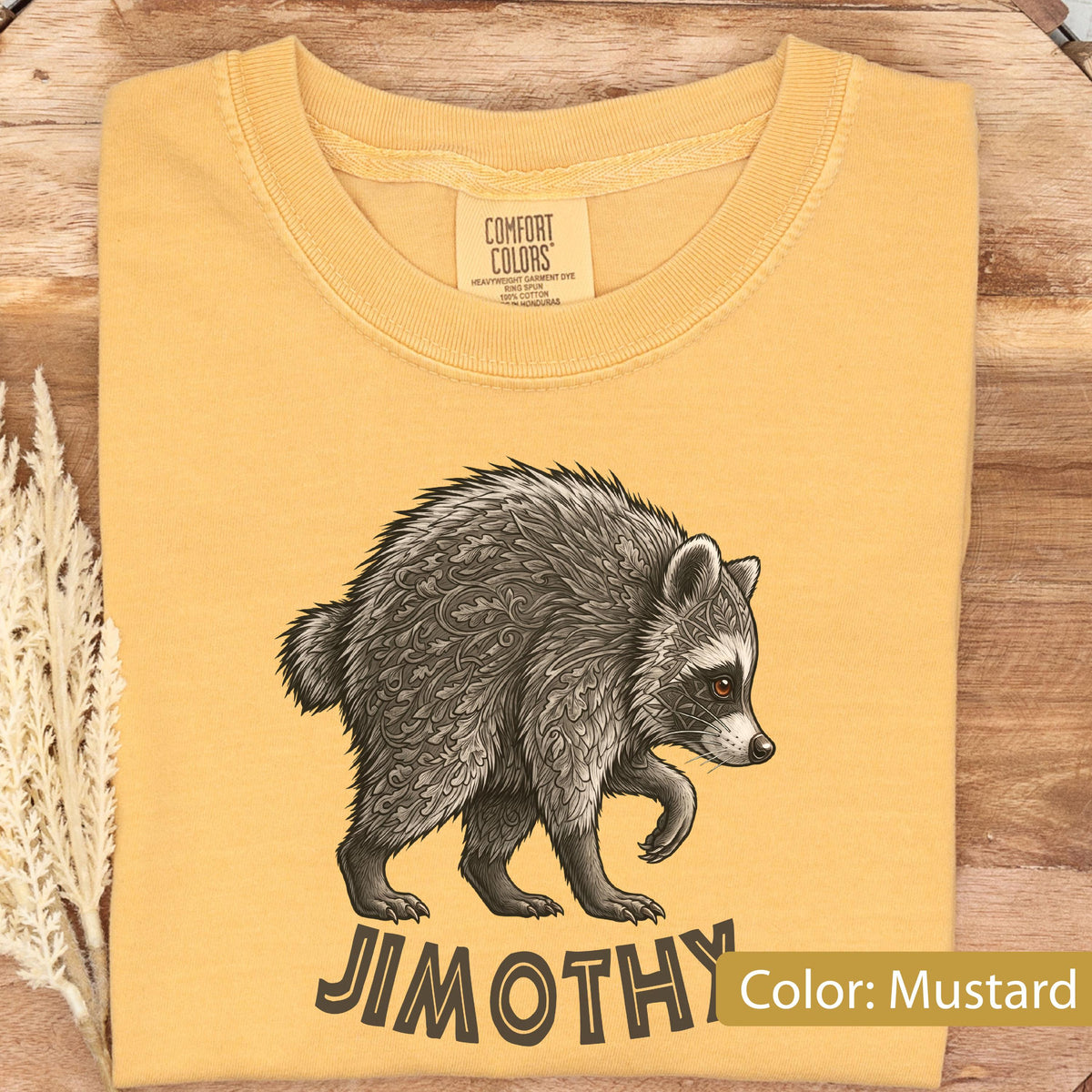 May include: A mustard yellow t-shirt featuring a graphic illustration of a raccoon with intricate leaf and geometric patterns on its fur. The text JIMOTHY is printed in a bold, dark font below the raccoon. The shirt has a Comfort Colors label.