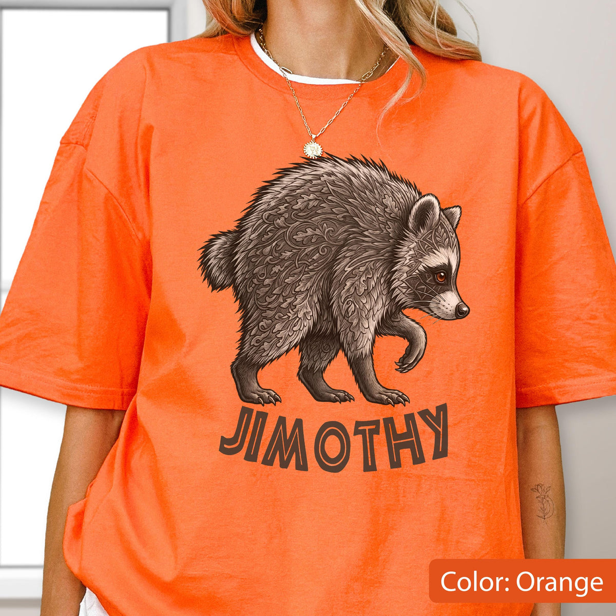 May include: A bright orange short-sleeve t-shirt featuring a graphic of a raccoon with intricate leaf-like patterns in its fur. Below the raccoon, the text JIMOTHY is printed in a dark brown, bold, sans-serif font. The shirt has a crew neckline.