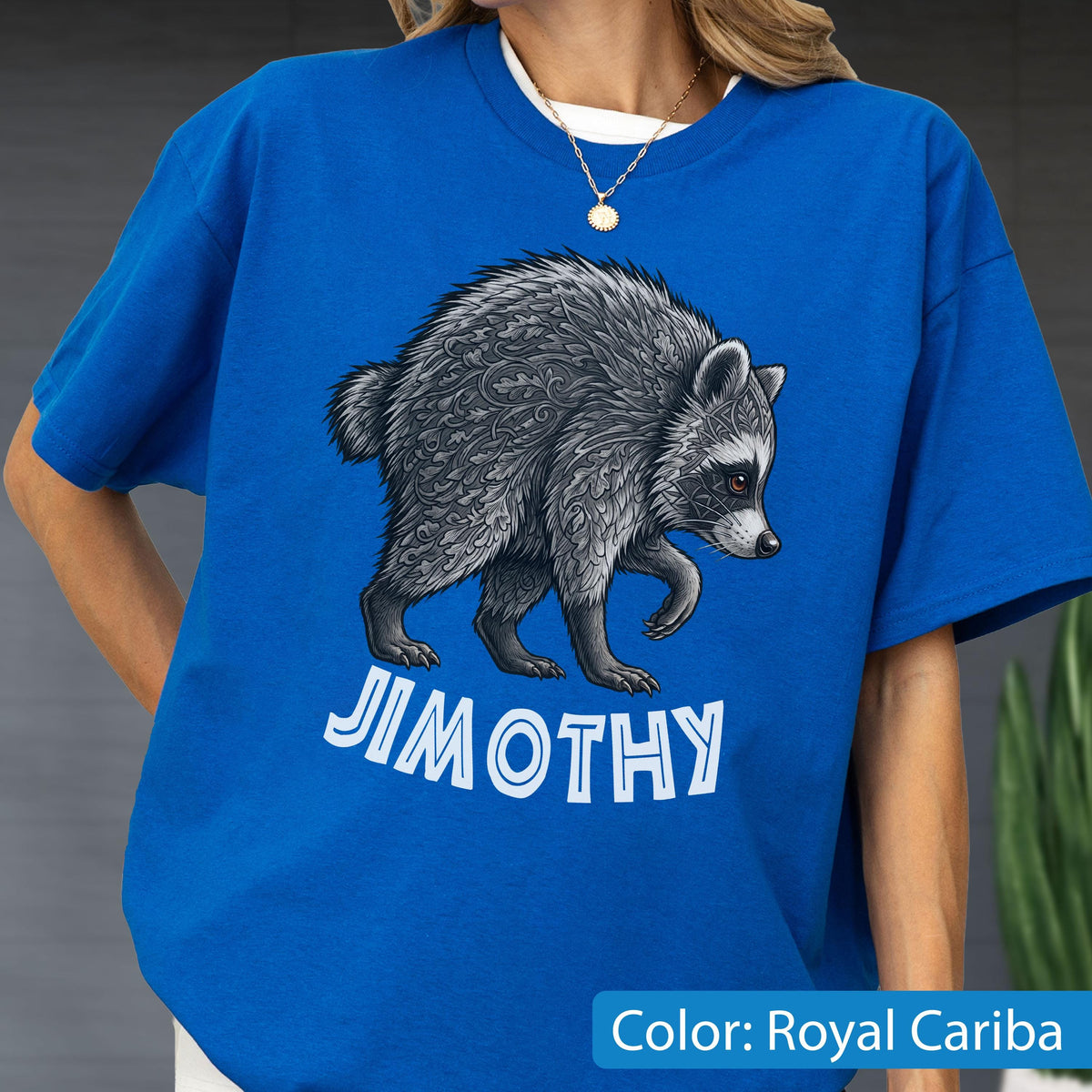 May include: A royal blue short-sleeve t-shirt featuring a graphic of a grey raccoon with intricate leaf-like patterns in its fur. Below the raccoon, the text JIMOTHY is printed in bold, white, capital letters. The shirt has a crew neckline.