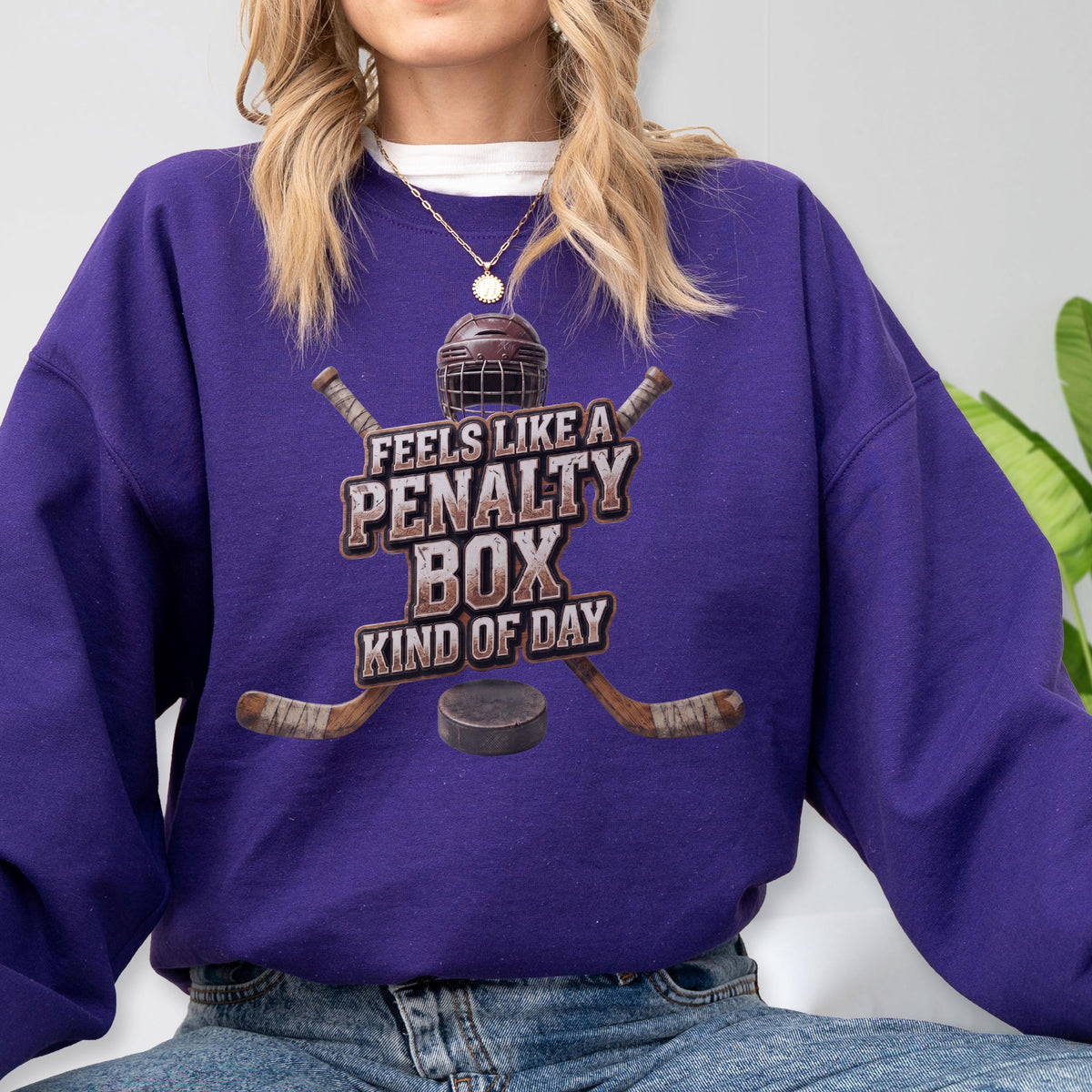May include: A purple long-sleeve sweatshirt featuring a graphic of a hockey helmet, two crossed hockey sticks, and a puck. The text reads FEELS LIKE A PENALTY BOX KIND OF DAY. The design is centered on the chest of the garment.