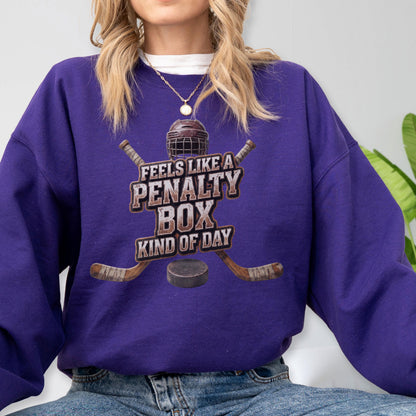 May include: A purple long-sleeve sweatshirt featuring a graphic of a hockey helmet, two crossed hockey sticks, and a puck. The text reads FEELS LIKE A PENALTY BOX KIND OF DAY. The design is centered on the chest of the garment.