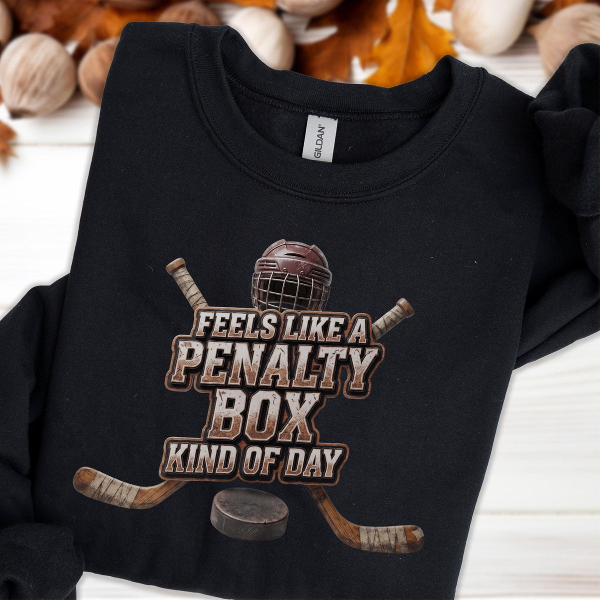 May include: A black crewneck sweatshirt featuring a graphic of a hockey helmet, two crossed hockey sticks, and a puck. The text reads "FEELS LIKE A PENALTY BOX KIND OF DAY" in a distressed, weathered font style centered on the chest of the garment.