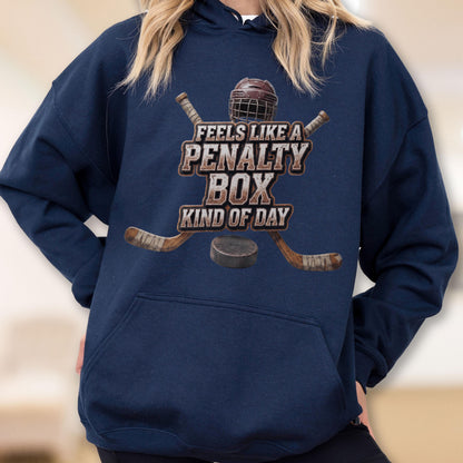 May include: A navy blue hooded sweatshirt featuring a graphic print of two crossed hockey sticks, a hockey helmet, and a puck. The text reads, "FEELS LIKE A PENALTY BOX KIND OF DAY" in a distressed, block-style font centered on the chest.
