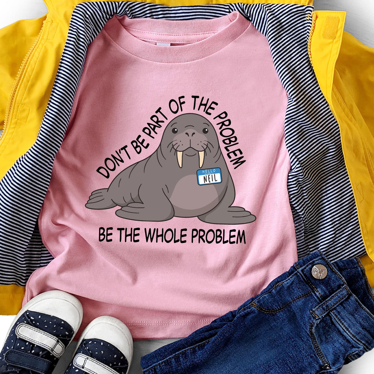 May include: A light pink t-shirt featuring a graphic illustration of a grey walrus wearing a blue name tag that says "HELLO NEIL". The text "DON'T BE PART OF THE PROBLEM" is arched above the walrus, and "BE THE WHOLE PROBLEM" is printed below.