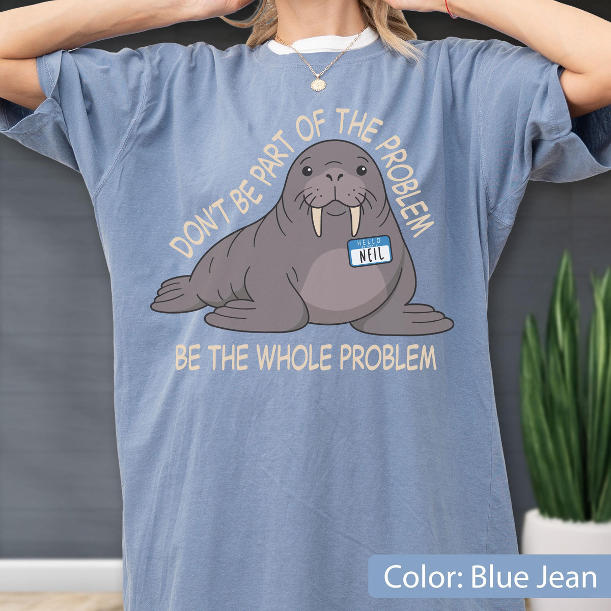 May include: A blue jean colored t-shirt featuring a graphic of a walrus wearing a name tag that reads NEIL. The text above the walrus reads DON'T BE PART OF THE PROBLEM and the text below the walrus reads BE THE WHOLE PROBLEM in light yellow.