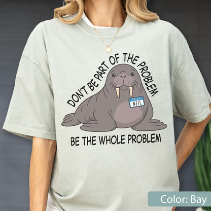 May include: A light sage green t-shirt featuring a graphic of a grey walrus wearing a blue name tag that reads "HELLO NEIL". The text above the walrus reads "DON'T BE PART OF THE PROBLEM" and the text below reads "BE THE WHOLE PROBLEM" in black.