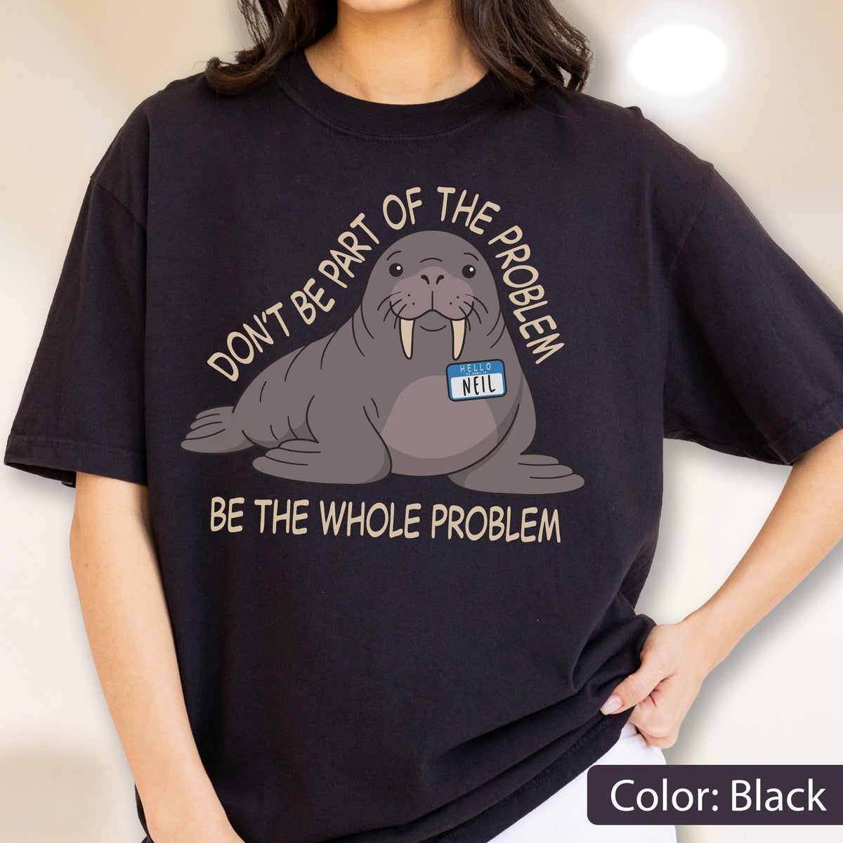 May include: A black t-shirt featuring a graphic of a grey walrus wearing a blue name tag that reads "HELLO NEIL". The text "DON'T BE PART OF THE PROBLEM" is arched above the walrus, and "BE THE WHOLE PROBLEM" is printed in beige below it.