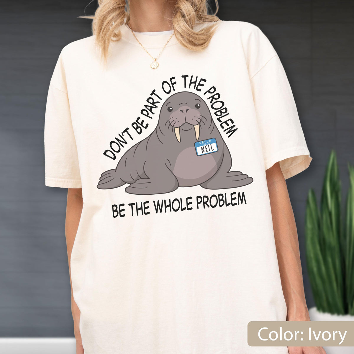 May include: An ivory t-shirt featuring a graphic of a grey walrus wearing a blue name tag that reads "HELLO NEIL". The text above the walrus reads "DON'T BE PART OF THE PROBLEM" and the text below the walrus reads "BE THE WHOLE PROBLEM".