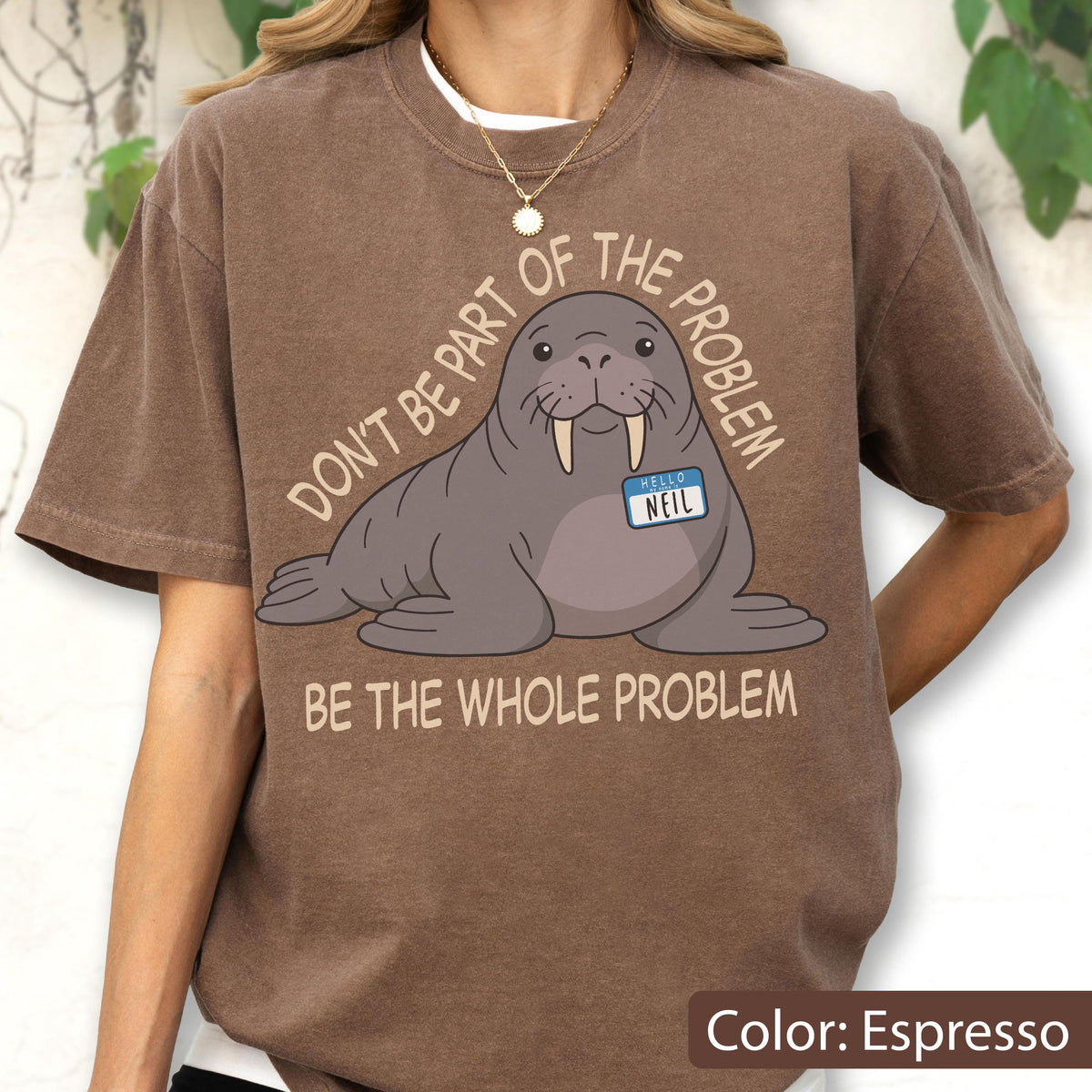 May include: An espresso-colored t-shirt featuring a cartoon walrus with a name tag that reads "HELLO NEIL". The text above the walrus says "DON'T BE PART OF THE PROBLEM" and the text below the walrus says "BE THE WHOLE PROBLEM" in light tan.