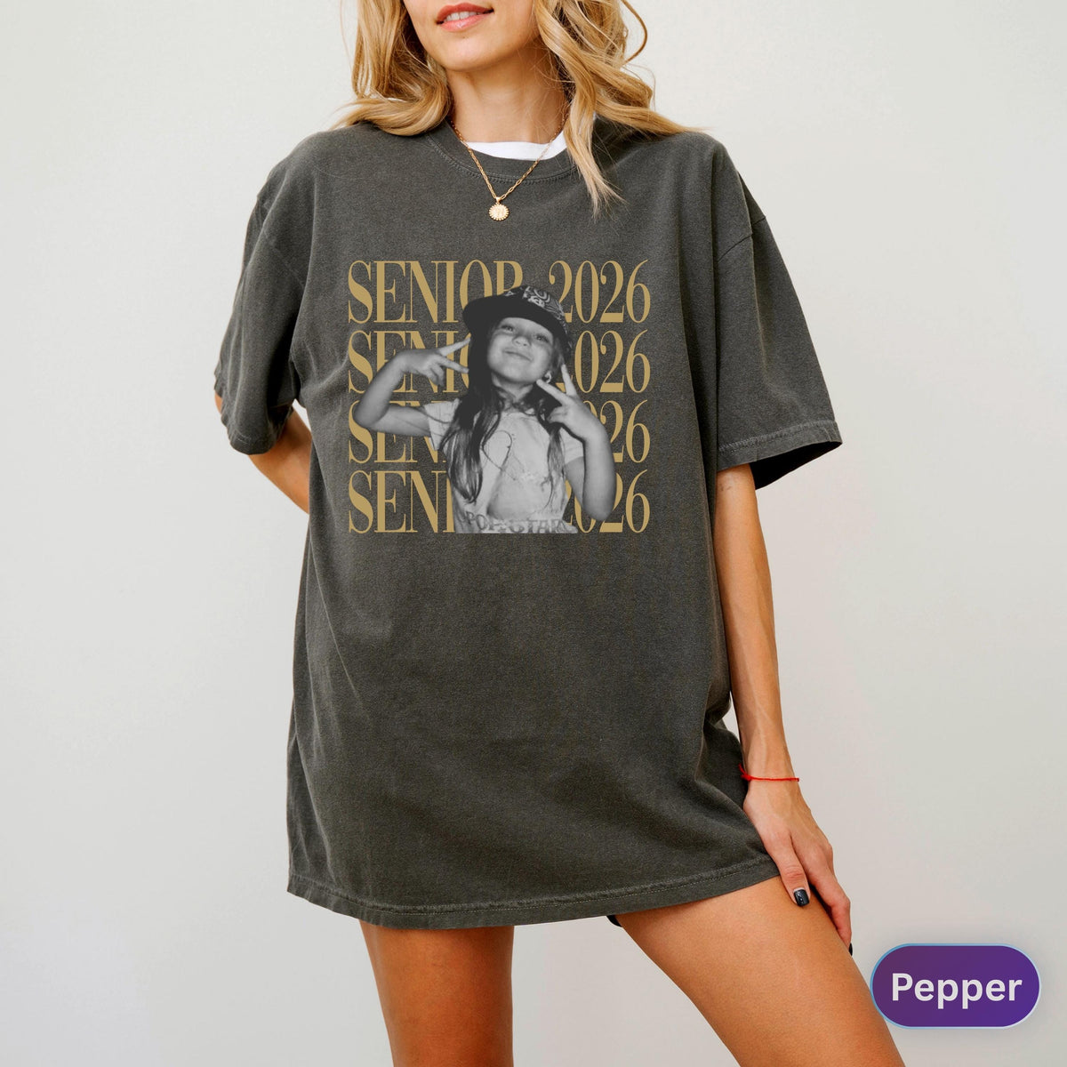 May include: A charcoal gray short-sleeve t-shirt featuring a central graphic of a person wearing a cap and making a peace sign. The text SENIOR 2026 is printed in a large, repeating gold font behind the central black and white image.