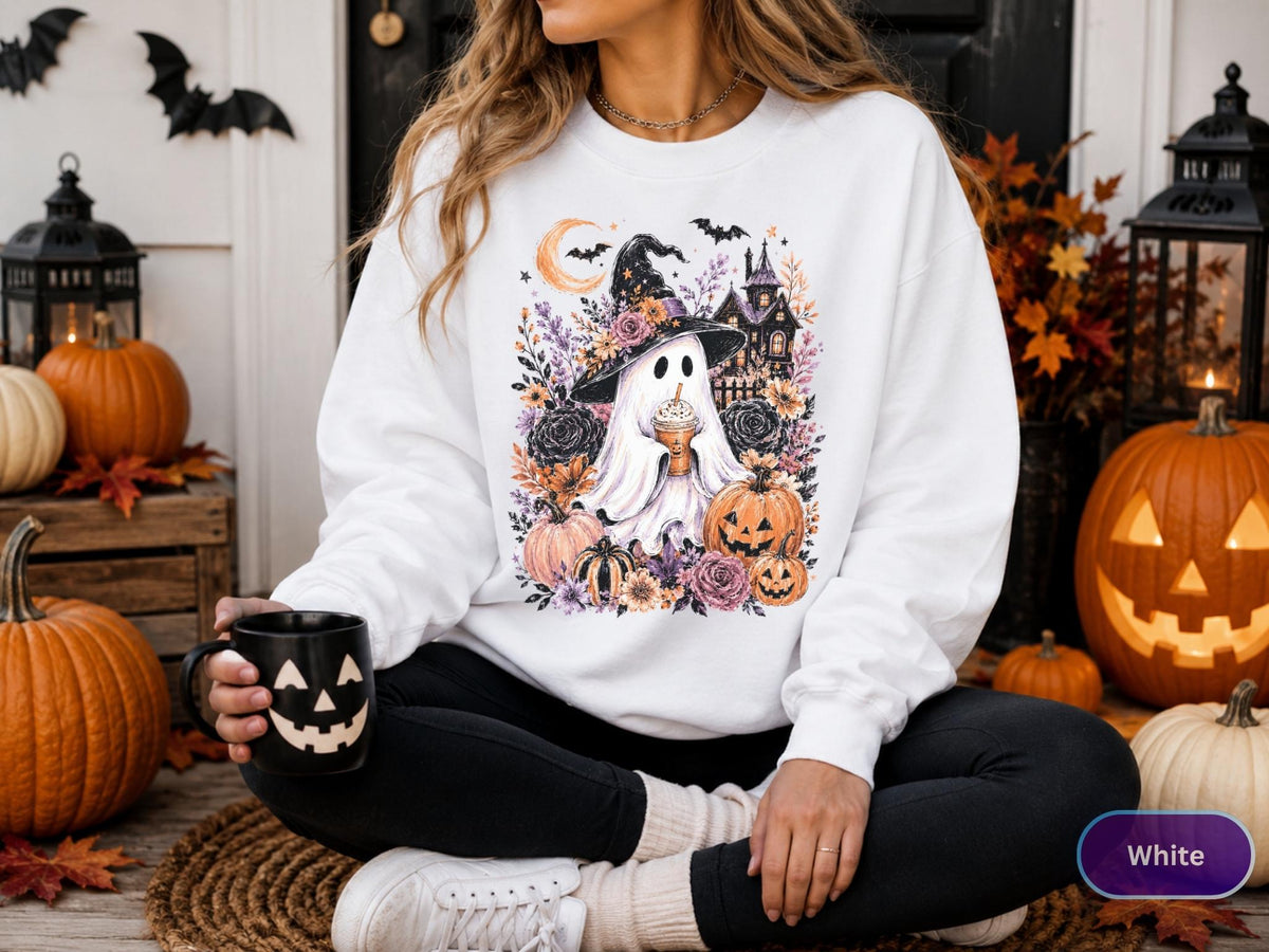 May include: A white long-sleeve sweatshirt featuring a graphic print of a ghost wearing a witch hat and holding a coffee cup, surrounded by pumpkins, flowers, a crescent moon, bats, and a haunted house illustration in shades of orange and purple.