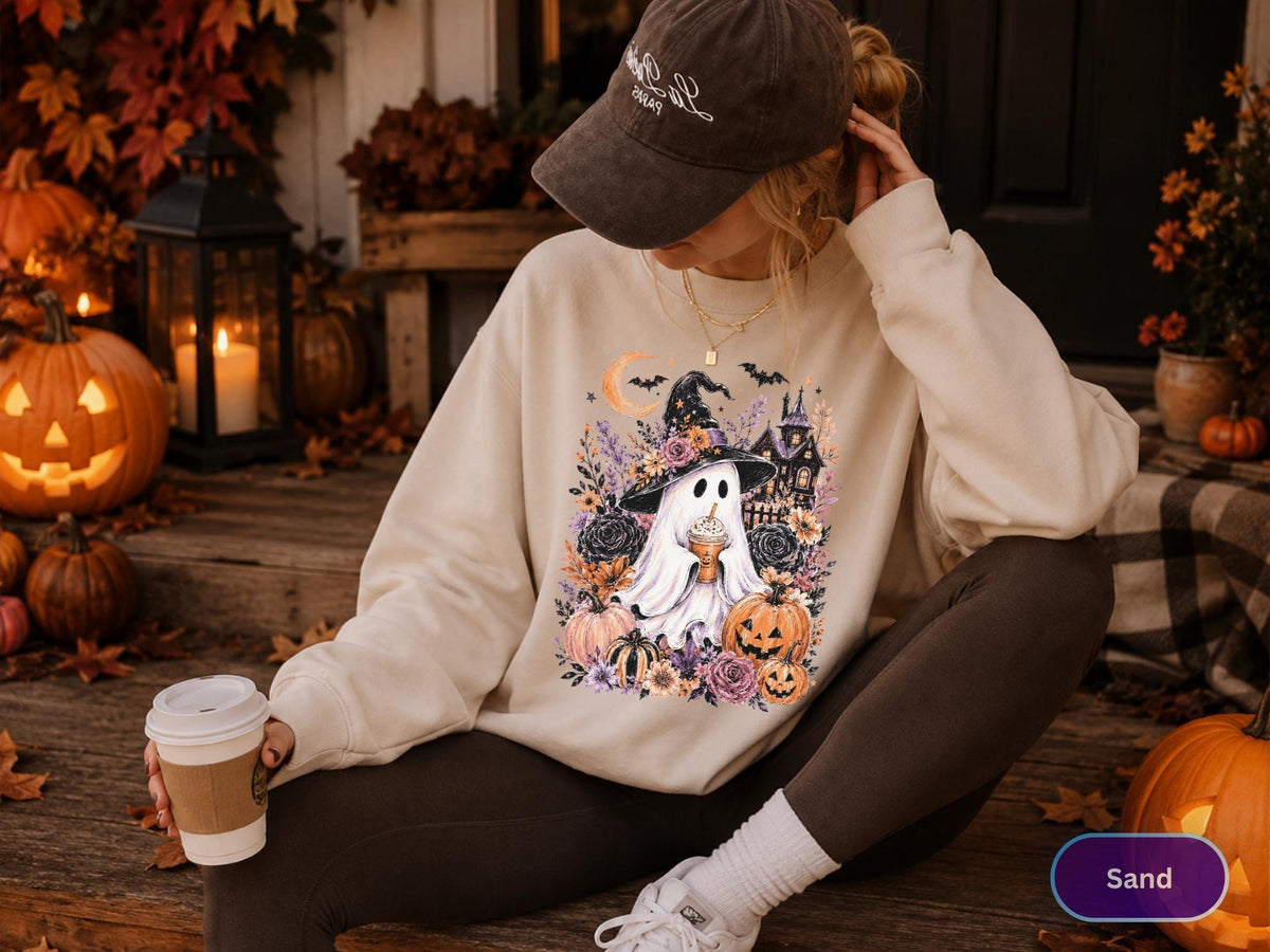 May include: A sand-colored crewneck sweatshirt featuring a graphic print of a ghost wearing a witch hat and holding a coffee cup, surrounded by pumpkins, flowers, and a haunted house. The sweatshirt is paired with dark leggings and a baseball cap.