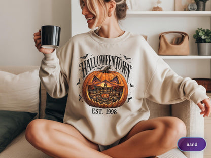 May include: A sand-colored crewneck sweatshirt featuring a large graphic print of a carved orange pumpkin with a detailed town scene inside. The text HALLOWEENTOWN is arched above the pumpkin, with EST. 1998 printed below it, surrounded by small bats.