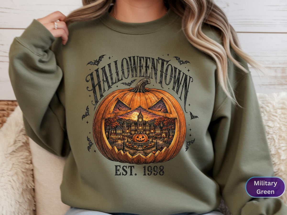 May include: A military green crewneck sweatshirt featuring a graphic of a large carved pumpkin with a detailed town scene inside. The text HALLOWEENTOWN is printed above the pumpkin, and EST. 1998 is printed below, surrounded by small black bats.