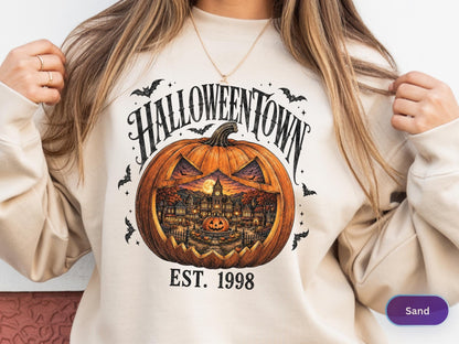 May include: A sand-colored crewneck sweatshirt featuring a graphic print of a large orange jack-o'-lantern with a detailed town scene inside. The text reads HALLOWEENTOWN above the pumpkin and EST. 1998 below, surrounded by small black bats.