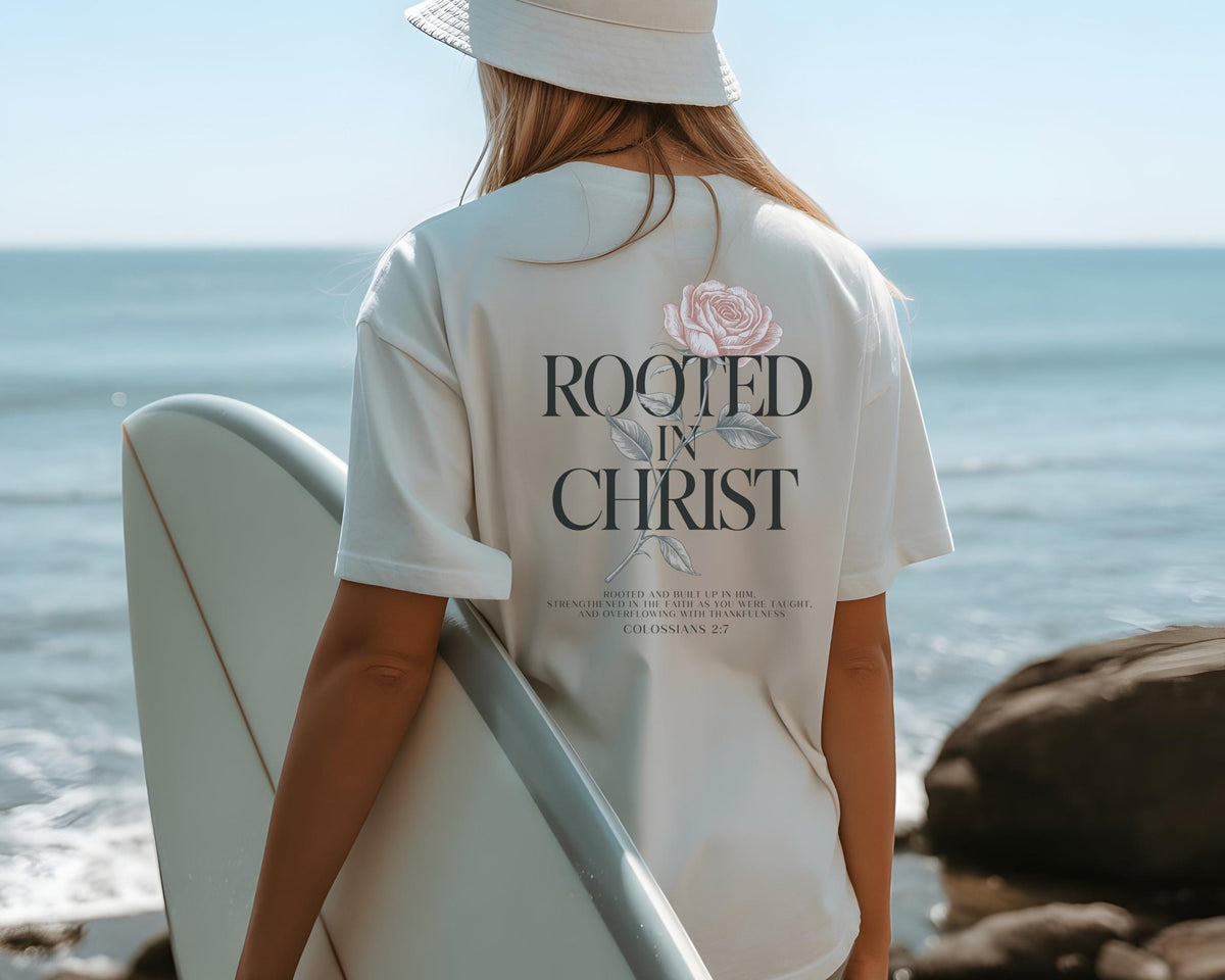 May include: A cream-colored short-sleeve t-shirt featuring a graphic on the back with a pink rose illustration, the text "ROOTED IN CHRIST," and the scripture reference "COLOSSIANS 2:7" printed in a dark serif font above a small block of text.