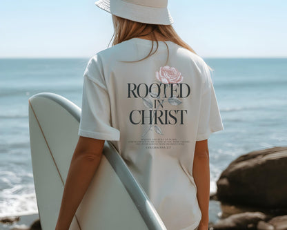 May include: A cream-colored short-sleeve t-shirt featuring a graphic on the back with a pink rose illustration, the text "ROOTED IN CHRIST," and the scripture reference "COLOSSIANS 2:7" printed in a dark serif font above a small block of text.