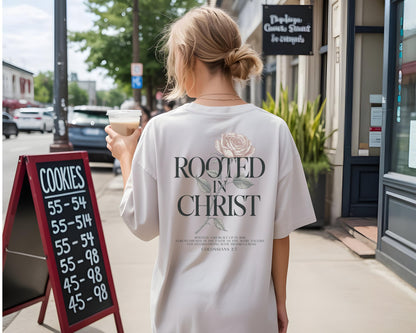 May include: A light beige short-sleeved t-shirt featuring a graphic on the back with a pale pink rose illustration and the black text "ROOTED IN CHRIST" above smaller scripture text from Colossians 2:7, displayed on a person from behind.