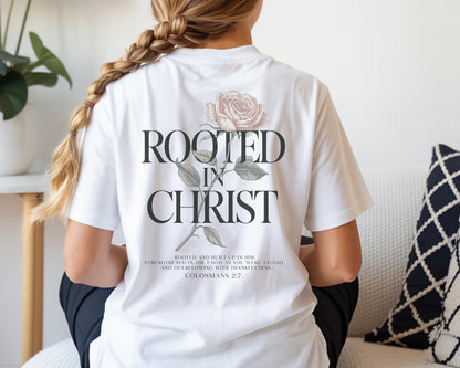 May include: A white t-shirt featuring a back graphic of a rose with the text "ROOTED IN CHRIST" in large dark letters. Below the text, smaller print reads "ROOTED AND BUILT UP IN HIM, STRENGTHENED IN THE FAITH AS YOU WERE TAUGHT, AND OVERFLOWING WITH THANKFULNESS COLOSSIANS 2:7".