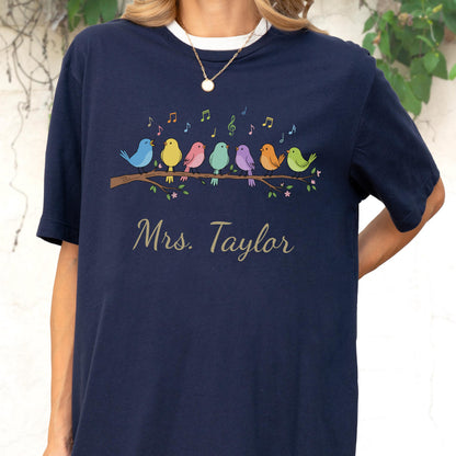 May include: A navy blue t-shirt featuring a graphic of seven colorful birds perched on a brown branch with musical notes floating above them. Below the birds, the text "Mrs. Taylor" is printed in a light-colored, cursive script font.