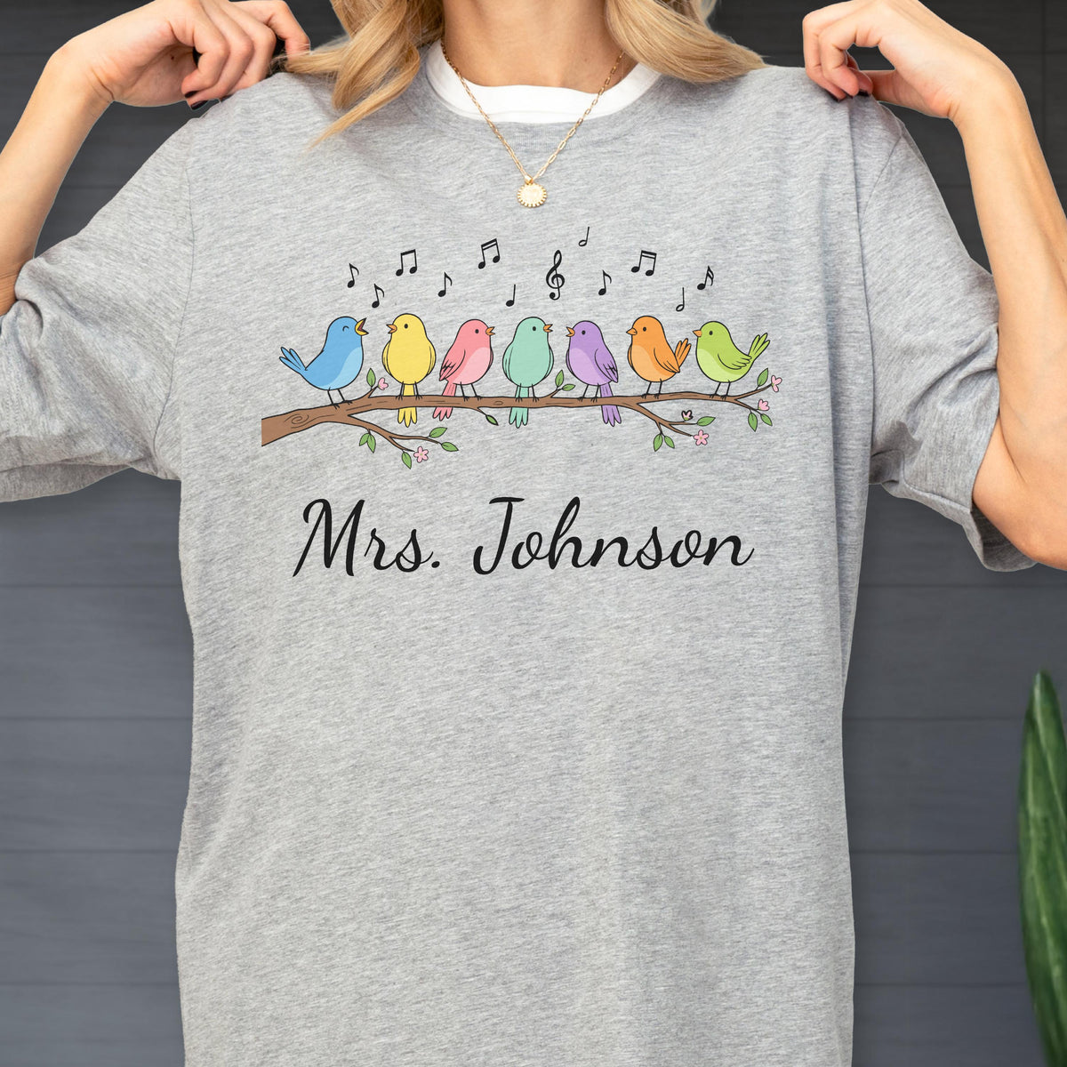 May include: A heather gray t-shirt featuring a graphic of seven colorful birds perched on a brown tree branch with musical notes floating above them. The text "Mrs. Johnson" is printed in black cursive script below the illustrated birds.