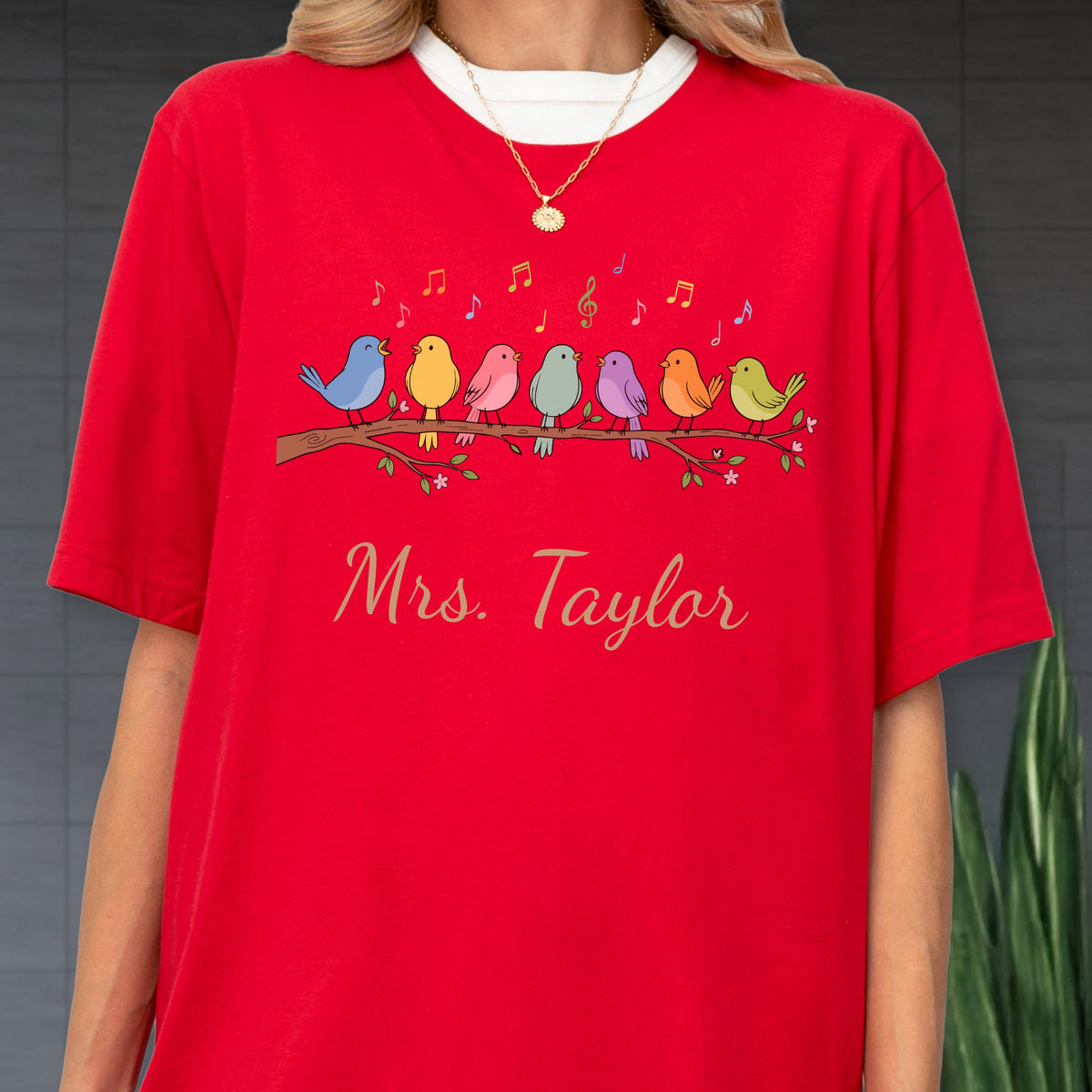 May include: A bright red short-sleeved t-shirt featuring a graphic of seven colorful birds perched on a brown branch with musical notes above them. Below the birds, the text Mrs. Taylor is printed in a cursive gold font. A gold necklace is worn.