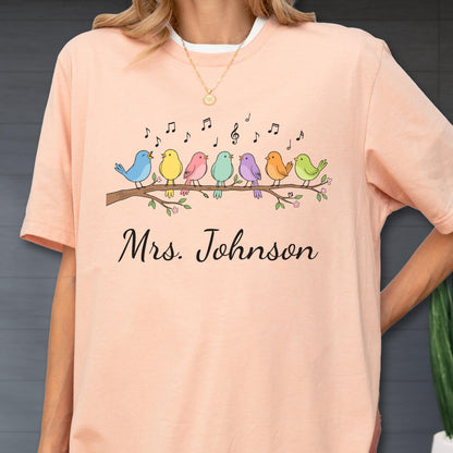 May include: A peach-colored t-shirt featuring a graphic of seven colorful birds perched on a brown branch with musical notes floating above them. Below the birds, the text Mrs. Johnson is printed in a black cursive script font on the front.