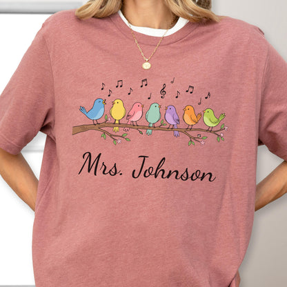 May include: A dusty rose short-sleeve t-shirt featuring a graphic of seven colorful birds perched on a branch with musical notes above them. Below the birds, the text "Mrs. Johnson" is printed in a black, cursive, script-style font.