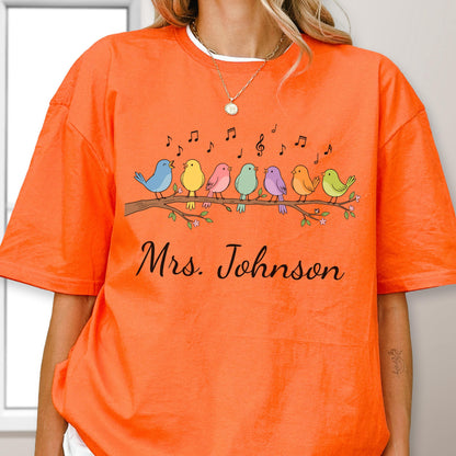 May include: An orange short-sleeved t-shirt featuring a graphic of seven colorful birds perched on a brown branch with musical notes floating above them. Below the birds, the text Mrs. Johnson is printed in a black cursive script font.