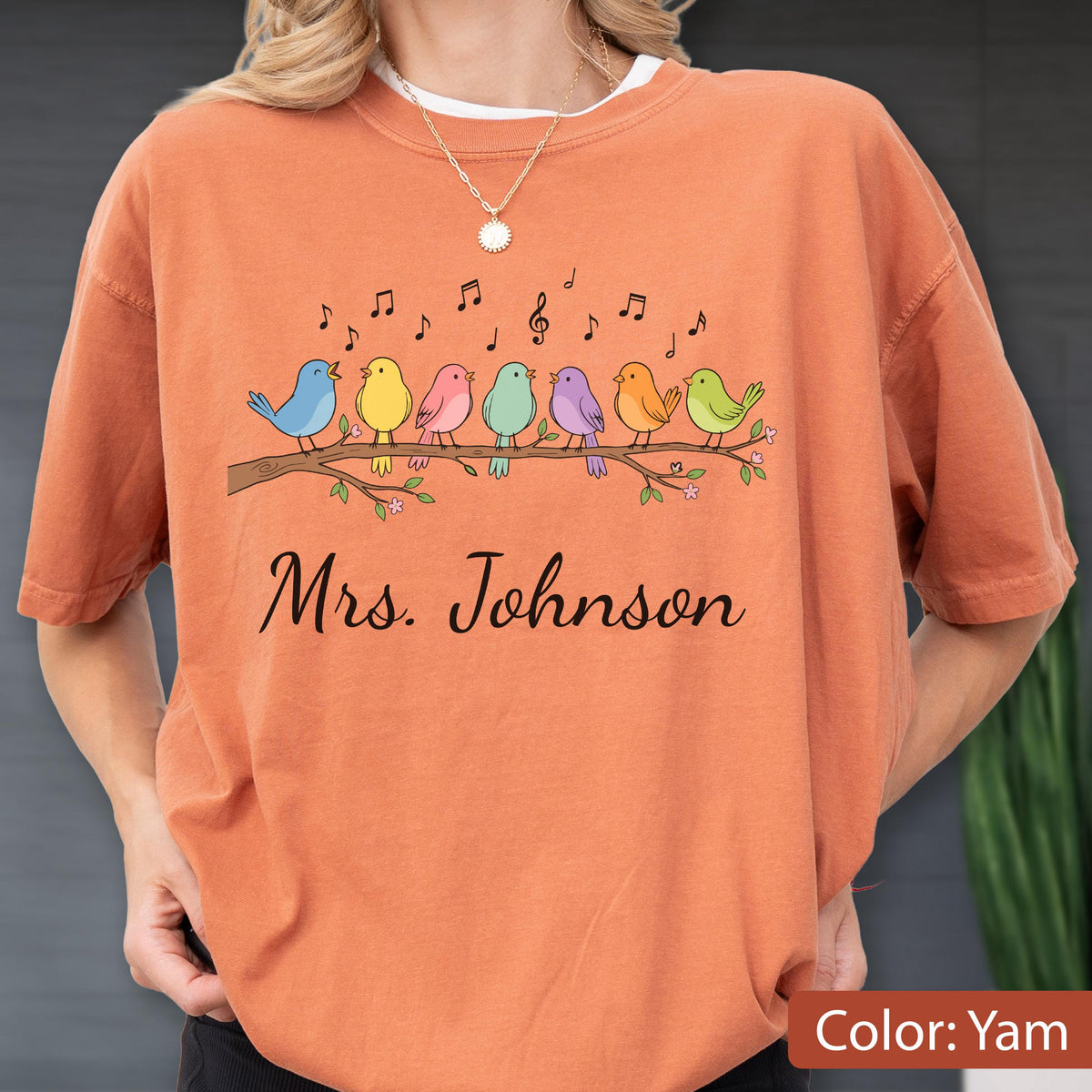 May include: A yam-colored t-shirt featuring a graphic of seven colorful birds perched on a branch with musical notes above them. The text "Mrs. Johnson" is printed in black script below the birds. The shirt has a crew neckline and short sleeves.