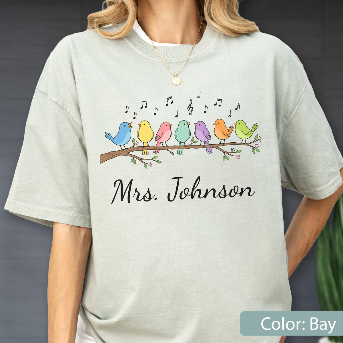 May include: A light sage green short-sleeve t-shirt featuring a graphic of seven colorful birds perched on a brown branch with musical notes above them. The text "Mrs. Johnson" is printed in black cursive script below the bird illustration.