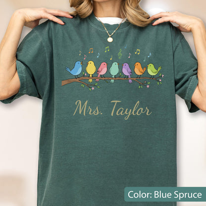 May include: A blue spruce t-shirt featuring a graphic of seven colorful birds perched on a brown branch with musical notes above them. Below the branch, the text Mrs. Taylor is printed in a light-colored cursive script font.