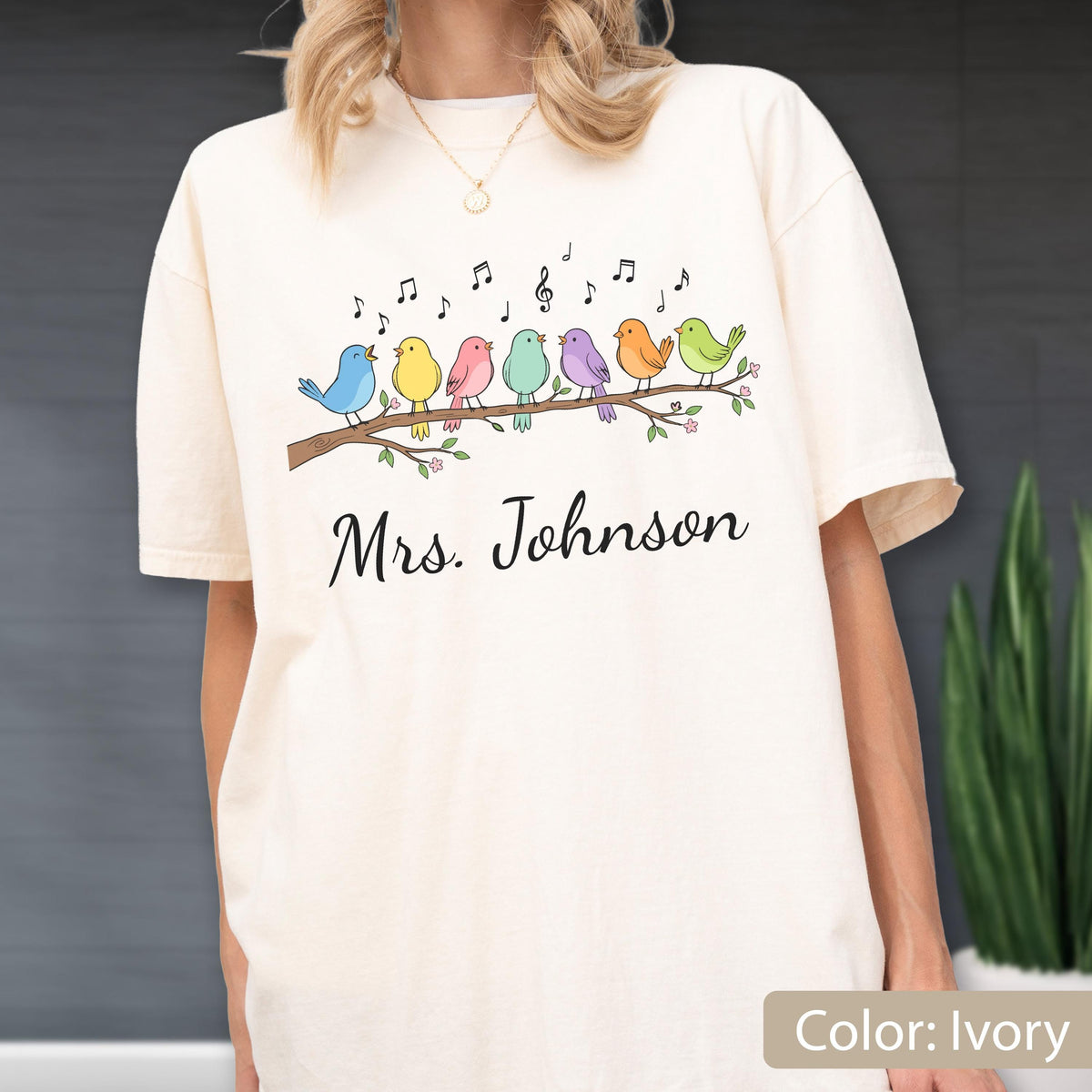May include: An ivory t-shirt featuring a graphic of seven colorful birds perched on a brown tree branch with musical notes floating above them. Below the branch, the text Mrs. Johnson is printed in a black cursive script font.
