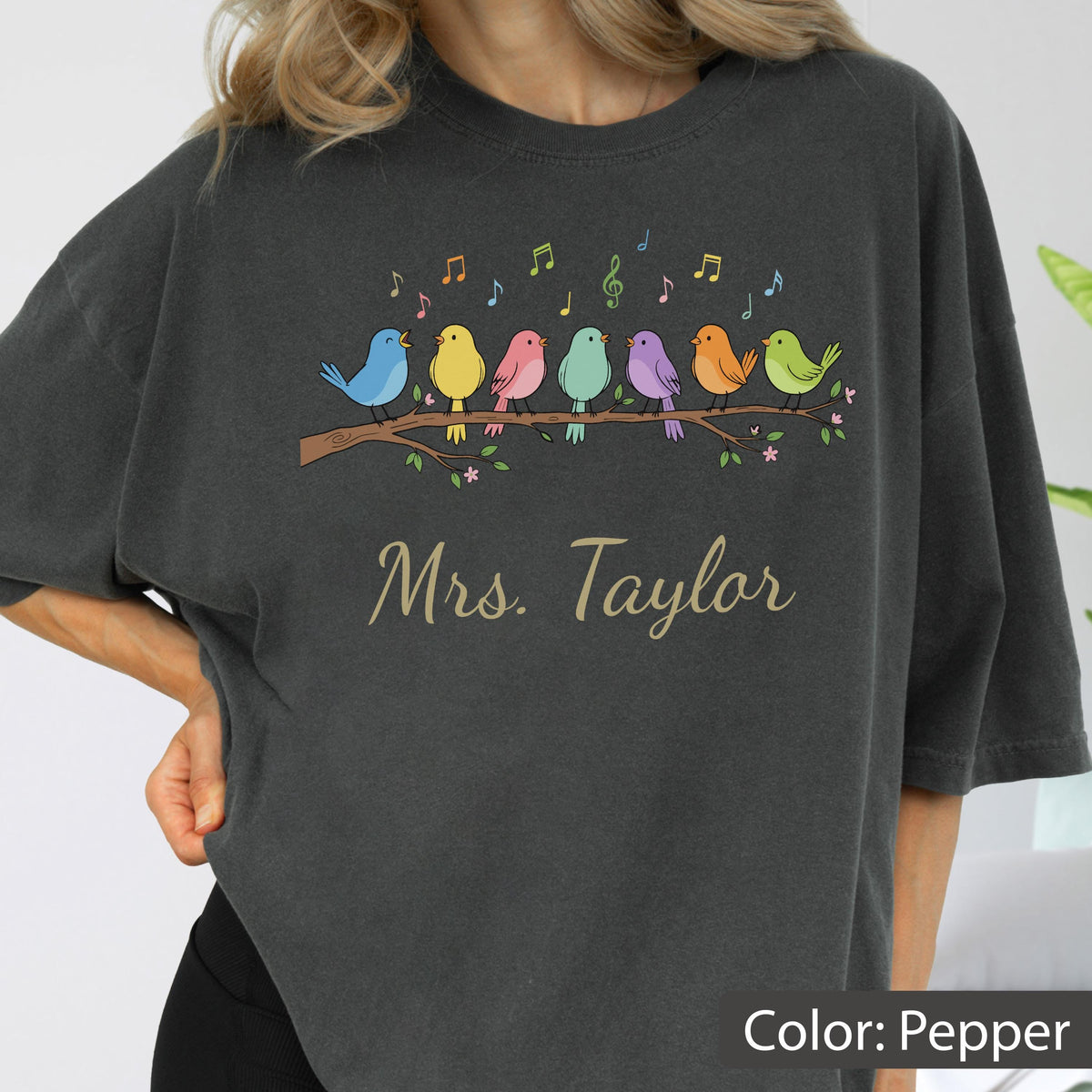 May include: A charcoal gray t-shirt featuring a graphic of seven colorful birds perched on a brown tree branch with musical notes above them. The text "Mrs. Taylor" is printed in a light tan script font below the birds on the front of the shirt.