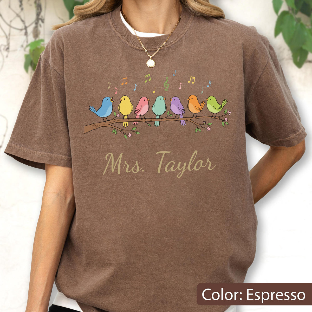 May include: An espresso-colored t-shirt featuring a graphic of seven colorful birds perched on a branch with musical notes floating above them. Below the birds, the text Mrs. Taylor is printed in a cursive font. The shirt has a crew neckline.