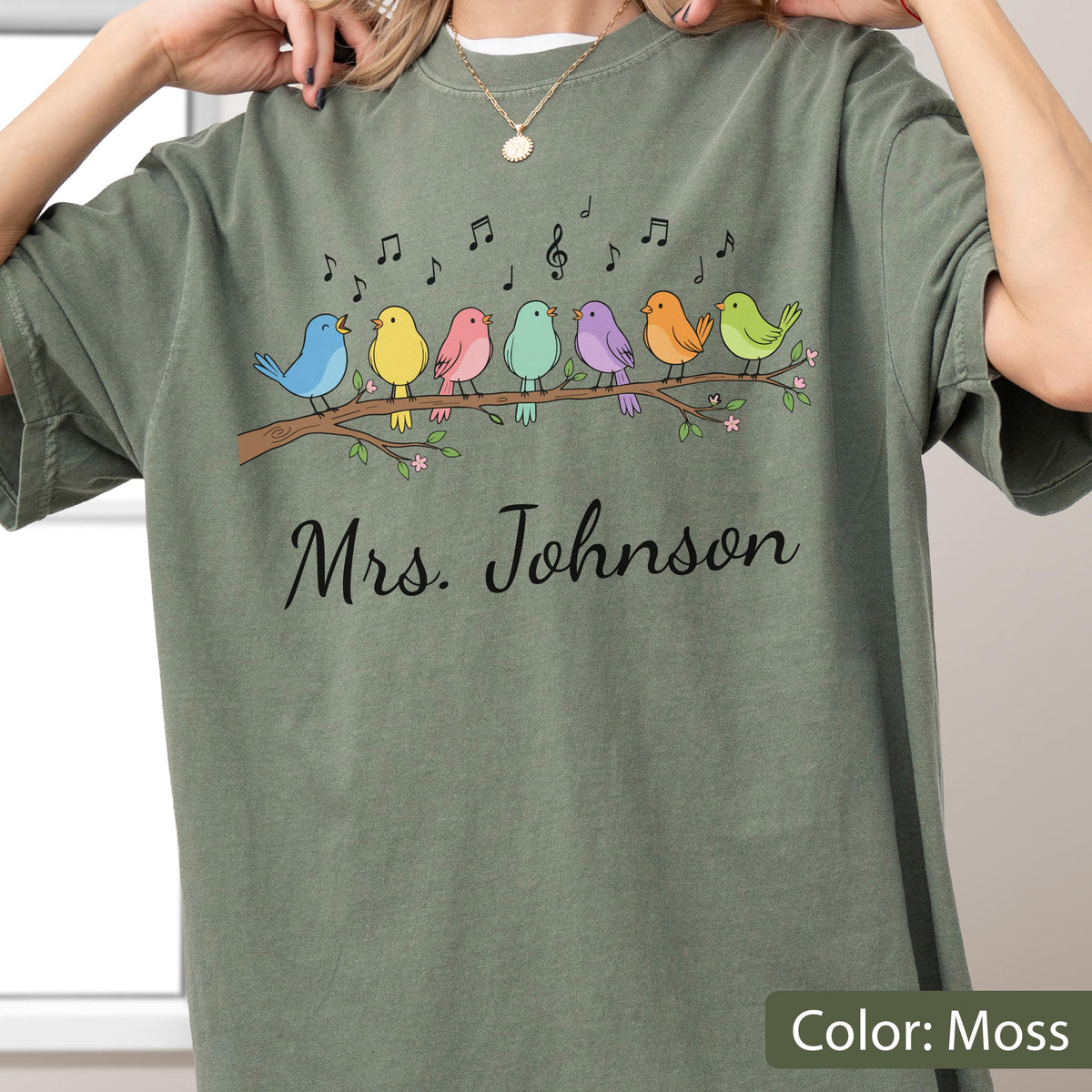 May include: A moss green t-shirt featuring a graphic of seven colorful birds perched on a brown tree branch with musical notes floating above them. Below the birds, the text Mrs. Johnson is printed in a black cursive script font.