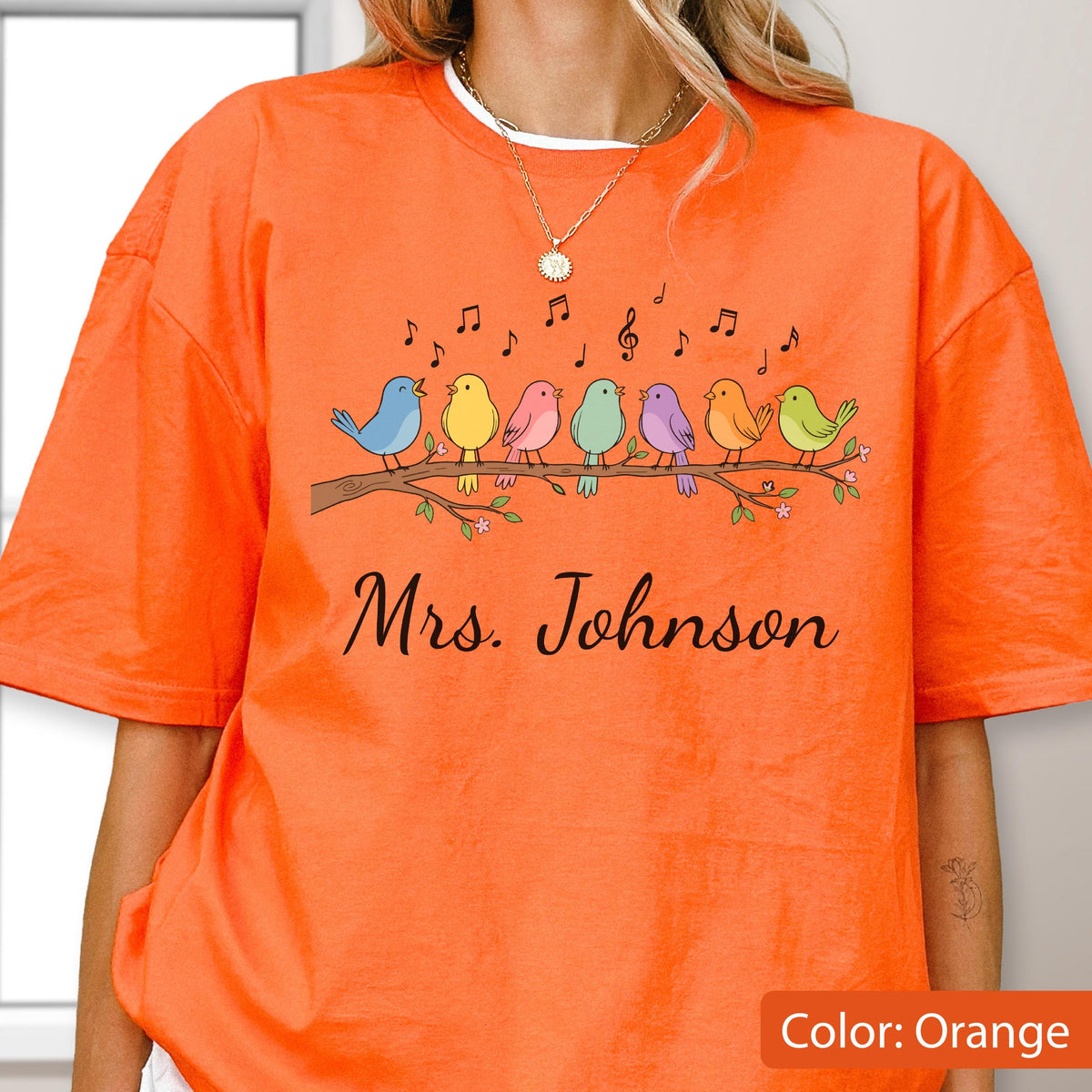 May include: An orange short-sleeved t-shirt featuring a graphic of seven colorful birds perched on a brown branch with musical notes floating above them. Below the birds, the text Mrs. Johnson is printed in a black cursive script font.