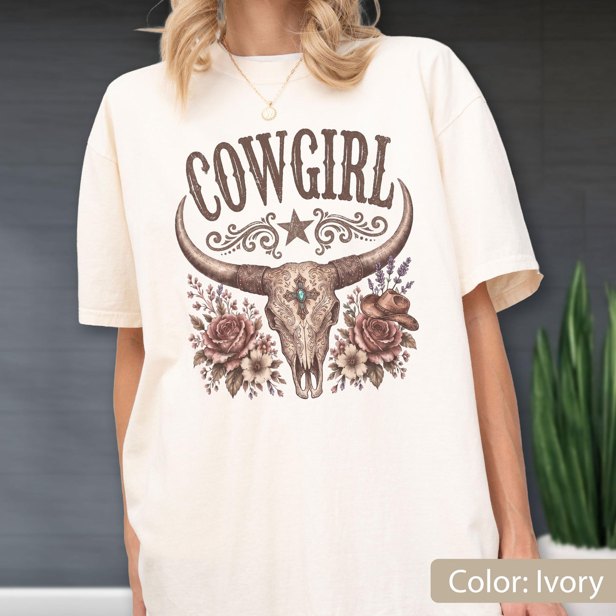May include: An ivory short-sleeve t-shirt featuring a graphic print of a cow skull with long horns, a central turquoise stone, and floral accents. The text COWGIRL is printed in a brown western-style font above the skull, with a star and swirls.