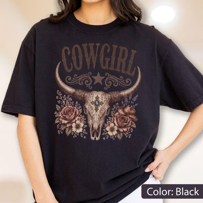 May include: A black t-shirt featuring a graphic print of a longhorn skull with ornate filigree patterns, a central turquoise stone, and a small cowboy hat. The design includes floral accents and the text COWGIRL in a decorative brown font.