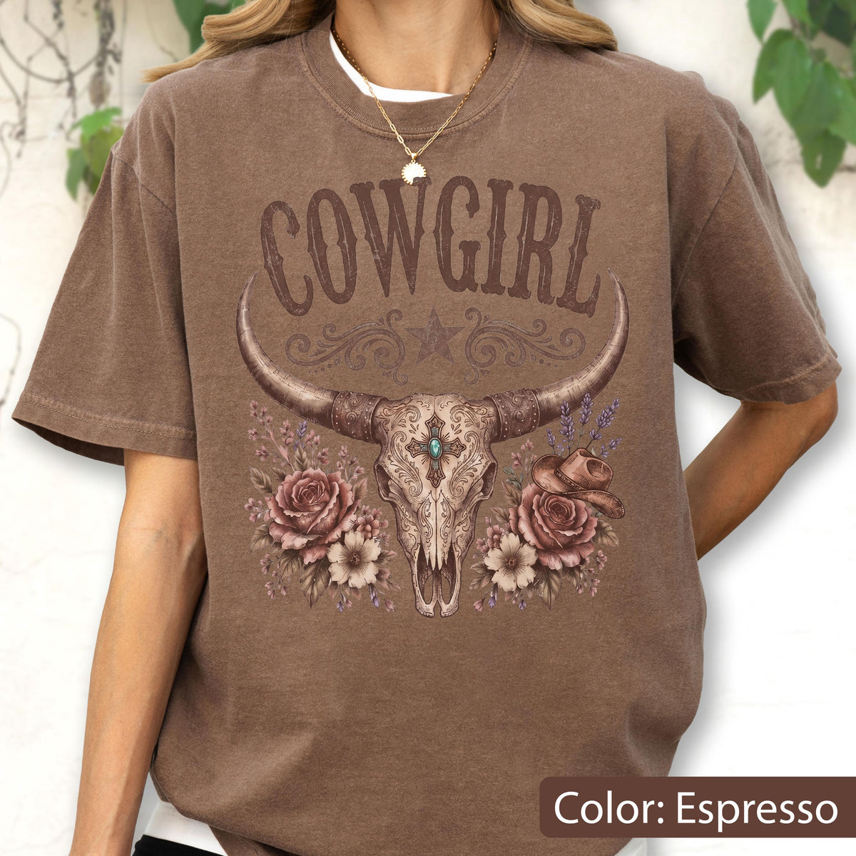 May include: An espresso-colored t-shirt featuring a graphic print of a longhorn skull with ornate filigree, a central turquoise stone, roses, and a small cowboy hat. The text COWGIRL is printed in a decorative brown font above the skull design.